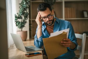 Stressed man sitting at a desk with a laptop and phone, holding a brown envelope, showing emotions of worry and concern about potential bad news or financial issues